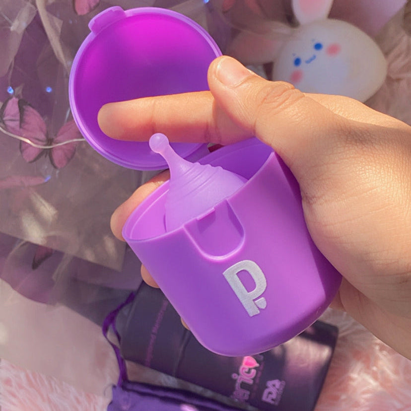 Purple baby bottle with a finger inserted into the nipple, held by a hand against a blurred background.